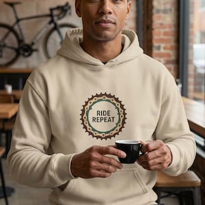 Puede incluir: Una sudadera con capucha de color beige que presenta un gráfico circular en el pecho. El diseño muestra un plato de bicicleta marrón con un anillo interior verde azulado y el texto RIDE REPEAT impreso en letras marrón oscuro en el centro.