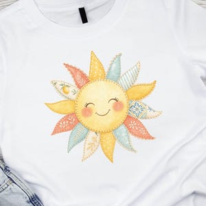May include: A white short-sleeved t-shirt featuring a central graphic of a smiling sun with a patchwork design. The sun's rays are illustrated in various pastel colors and patterns, including floral, polka dot, and lemon motifs, with stitched edges.