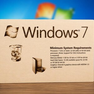 May include: A rectangular light-coloured wooden plaque engraved with the Windows 7 logo, a recycle bin icon, a folder icon, and the text Minimum System Requirements listing processor, memory, hard disk space, and graphics specifications in English.