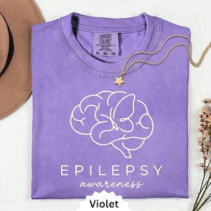 May include: A violet t-shirt featuring a white line-art graphic of a brain with a butterfly integrated into its shape. Below the graphic, the text reads EPILEPSY awareness. The shirt has a Comfort Colors label showing size XL and 100% cotton.