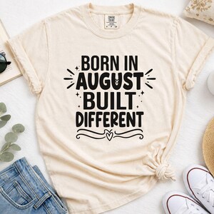 May include: A cream-colored short-sleeve t-shirt featuring black text that reads BORN IN AUGUST BUILT DIFFERENT. The design includes small star accents and a decorative flourish with a heart at the bottom. The shirt has a crew neckline.