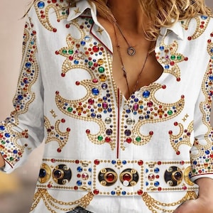 Op de afbeelding: Een wit overhemd met lange mouwen en knoopsluiting, voorzien van een printpatroon met gouden kettingen en kleurrijke imitatie-edelstenen in rood, blauw, groen en geel. Het ontwerp bevat een horizontale band in de taille als een riem.