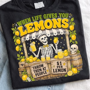 May include: A black t-shirt featuring a graphic of a skeleton at a lemonade stand with the text "WHEN LIFE GIVES YOU LEMONS" at the top and "THROW THEM AT PEOPLE" and "$1 PER LEMON" on the stand, surrounded by yellow lemons and green leaves.