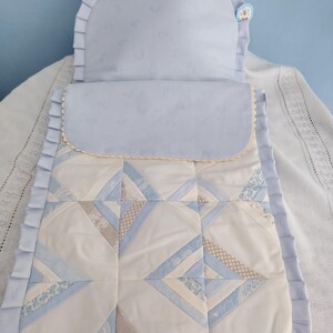 May include: A light blue and white quilted sleeping bag featuring a rounded hood with ruffled trim. The lower section displays a geometric patchwork pattern in shades of blue, white, and beige. A small circular tag is attached to the hood.