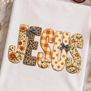 May include: A white t-shirt featuring the word JESUS in large, decorative letters. Each letter has a unique pattern including sunflowers, leopard print, orange gingham, polka dots, and pumpkins, accented with small fabric bows on the letters.