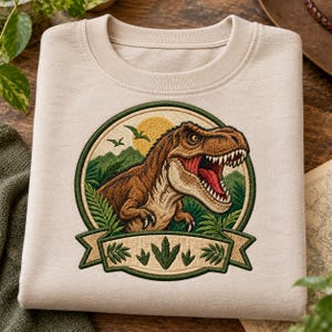 May include: A beige crewneck sweatshirt featuring a circular embroidered patch. The patch depicts a brown Tyrannosaurus Rex in a prehistoric landscape with green mountains, ferns, and a yellow sun. A banner at the bottom displays dinosaur footprints.