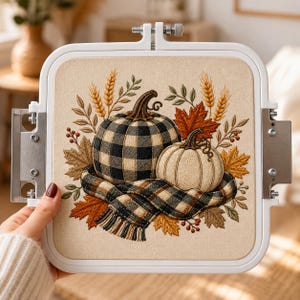 May include: A square embroidery hoop containing a fabric panel with a stitched design of two pumpkins, one with a black and white buffalo check pattern and one solid cream, wrapped in a matching plaid scarf, surrounded by autumn leaves and wheat.