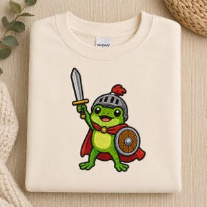May include: A cream-coloured crewneck sweatshirt featuring an embroidered graphic of a green frog dressed as a knight. The frog wears a silver helmet with a red plume, a red cape, and holds a silver sword in one hand and a round wooden shield in the other.