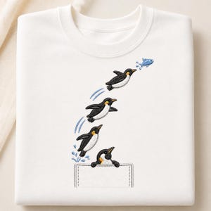 Op de afbeelding: Een wit sweatshirt met ronde hals voorzien van een borduursel van vier pinguïns. Eén pinguïn komt uit een gestikte zak, terwijl drie andere in een boog omhoog springen, waarbij de bovenste pinguïn een kleine blauwe vis achtervolgt met waterspatten.