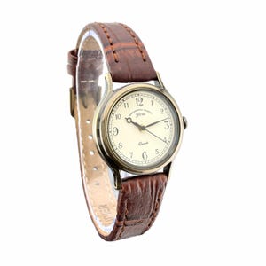 May include: A wristwatch with a round, antique-gold tone metal case and a cream-colored dial featuring black Arabic numerals. The dial displays the text Sentimental Graffiti you and Quartz. It has a brown, textured leather strap with stitching.