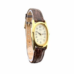 Può includere: Un orologio da polso color oro con cassa ovale e quadrante color crema con numeri arabi neri. Il quadrante riporta la scritta Isis e Citizen. È dotato di un cinturino in pelle marrone scuro con una texture a motivo coccodrillo.