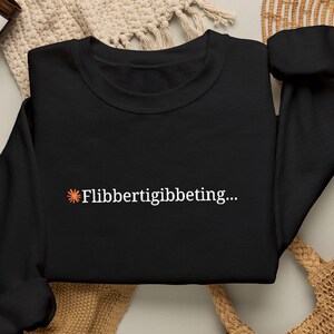 May include: A black long-sleeve sweatshirt featuring a small orange sun icon followed by the white text "Flibbertigibeting..." printed on the centre of the chest. The garment has a crew neckline and ribbed cuffs on the sleeves.