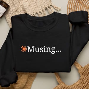 Puede incluir: Una sudadera negra de manga larga con un pequeño icono de estrella naranja seguido del texto blanco "Musing..." centrado en el pecho. La prenda tiene cuello redondo, mangas largas con puños acanalados y un dobladillo acanalado.