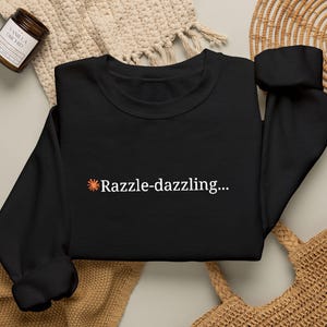 Puede incluir: Una sudadera negra de manga larga con un pequeño icono de estrella naranja seguido del texto blanco Razzle-dazzling... centrado en el pecho. La prenda tiene cuello redondo, mangas largas con puños acanalados y un dobladillo acanalado.