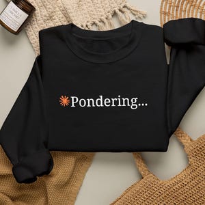 Puede incluir: Una sudadera negra de manga larga con un pequeño icono naranja en forma de sol seguido del texto blanco Pondering... centrado en el pecho. La prenda tiene cuello redondo y puños acanalados, ofreciendo un aspecto limpio y sencillo.