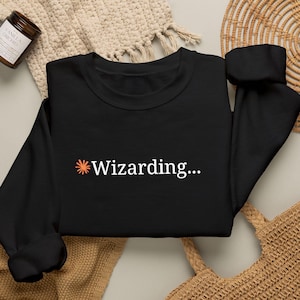 以下が含まれることがあります： 胸の中央に小さなオレンジ色のスターバーストアイコンと白い文字で「Wizarding...」とプリントされた、黒の長袖スウェットシャツ。クルーネックで、長袖の袖口はリブ編みになっています。