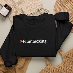 May include: A black long-sleeve sweatshirt featuring a small orange sunburst icon followed by the white text "Flummoxing..." centered on the chest. The garment has a crew neckline, long sleeves with ribbed cuffs, and a ribbed hemline.