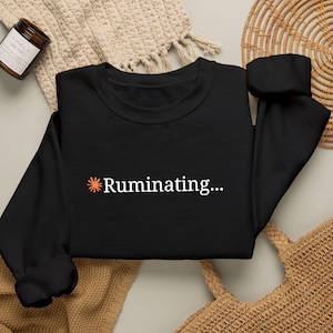 May include: A black long-sleeve sweatshirt featuring the text "Ruminating..." printed in white, accompanied by a small orange starburst icon to the left of the word. The garment has a crew neckline and ribbed cuffs on the sleeves.