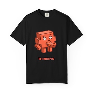 May include: A black short-sleeve t-shirt featuring a graphic of a blocky, orange, pixelated character with a thoughtful expression, resting its chin on its hand. Below the character, the word THINKING is printed in a matching orange pixelated font.