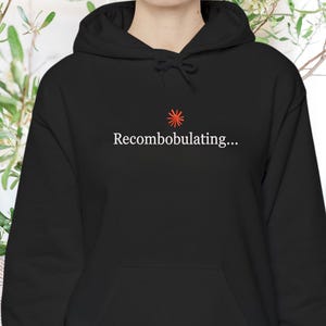 May include: A black hooded sweatshirt featuring a small red starburst icon centered above the white text "Recombobulating..." on the chest. The garment includes a drawstring hood, long sleeves with ribbed cuffs, and a front kangaroo pocket.