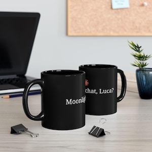 May include: Two black ceramic mugs featuring white text that reads Moonlight and chat, Luca? along with a small orange sun icon. The mugs have a standard cylindrical shape with a curved handle on the side, designed for holding hot or cold beverages.
