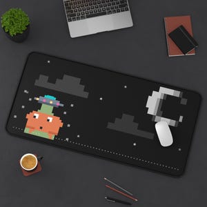 May include: A rectangular black desk mat featuring a pixel art design of an orange creature with a small UFO hovering above its head. The scene includes pixelated grey clouds, a white crescent moon, and small white stars scattered across the surface.