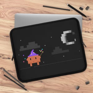 May include: A black laptop sleeve featuring a pixel art design of an orange creature with a purple pointed hat holding a small wand. The scene includes dark gray pixelated clouds, a white crescent moon, and small white stars against a black background.