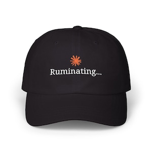 Può includere: Un cappellino da baseball nero con un'icona a stella arancione ricamata sopra il testo bianco "Ruminating..." centrato sul pannello frontale. Il cappellino presenta una corona strutturata, una visiera curva e un bottone sulla parte superiore.
