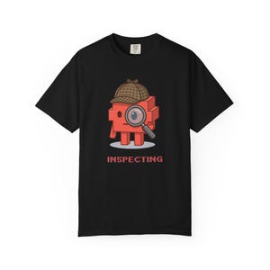 May include: A black short-sleeve t-shirt featuring a graphic of a red block-shaped character wearing a brown plaid deerstalker hat. The character holds a magnifying glass over one eye, and the word INSPECTING is printed in red below it.