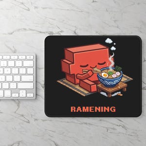 May include: A rectangular black mouse pad featuring a pixel art illustration of a red block-shaped character eating noodles from a blue bowl with chopsticks. The text RAMENING is printed in orange at the bottom of the mouse pad design.