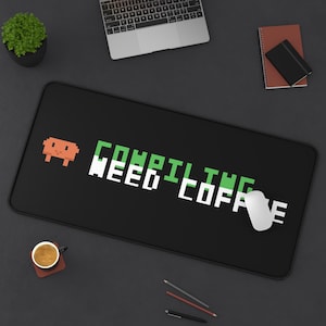 May include: A rectangular black desk mat featuring a pixelated orange alien icon on the left side. The text "COMPILING" appears in green pixelated letters above the white pixelated text "NEED COFFEE" centered on the mat surface.