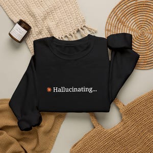 May include: A black long-sleeve sweatshirt featuring a small orange sun icon followed by the white text "Hallucinating..." printed on the center chest. The garment has a crew neckline, long sleeves with ribbed cuffs, and a ribbed hemline.