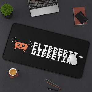 May include: A rectangular black desk mat featuring a pixelated orange creature graphic on the left and the white pixelated text "ELITEBEIT-LIEBETIN" on the right. The mat has a smooth surface and is positioned horizontally on a dark gray desk.