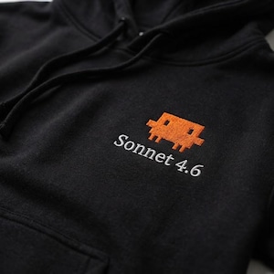 Könnte beinhalten: Ein schwarzer Kapuzenpullover mit einer gestickten orangefarbenen, verpixelten Kreatur über dem weißen Schriftzug Sonnet 4.6. Das Kleidungsstück verfügt über eine Kapuze mit Kordelzug und eine Kängurutasche auf der Vorderseite im Brustbereich.