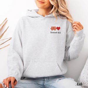May include: A light gray hooded sweatshirt featuring a small pixelated brown robot graphic next to a red pixelated heart on the left chest. Below the graphic, the text Sonnet 4.6 is printed in a simple black font. The hoodie has long sleeves.