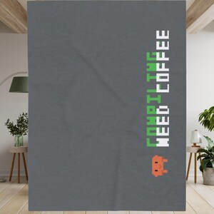 May include: A dark gray rectangular throw blanket featuring the vertical pixelated text "COMPILING NEED COFFEE" in green and white, accompanied by a small orange pixelated character graphic at the bottom right corner of the design.