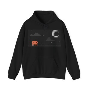 May include: A black hooded sweatshirt featuring a rectangular graphic print on the chest. The design displays a pixelated orange creature standing on a dotted line under a night sky with grey pixelated clouds, a crescent moon, and small white stars.