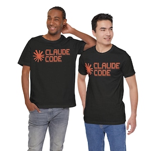 Puede incluir: Una camiseta negra de manga corta con un gráfico en el pecho que muestra un icono naranja de estallido solar junto al texto "CLAUDE CODE" en una fuente pixelada y cuadrada. El diseño está impreso en un color naranja vibrante sobre la tela.