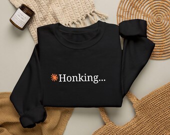 Op de afbeelding: Een zwart sweatshirt met lange mouwen en een kleine oranje ster-afbeelding, gevolgd door de witte tekst "Honking..." in het midden op de borst. Het kledingstuk heeft een ronde hals, lange mouwen met geribbelde boorden en een geribbelde zoom.