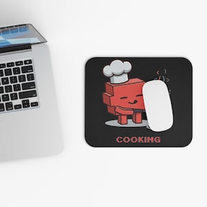 May include: A black rectangular mouse pad featuring a red pixelated block character wearing a white chef hat and holding a spatula. The graphic includes small white code brackets and the word COOKING in red pixelated text at the bottom.