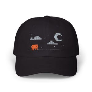 May include: A black baseball cap featuring an embroidered pixel art scene. The design includes a small orange square creature, two grey pixelated clouds, a grey pixelated crescent moon, and several small white stars on a dotted line.