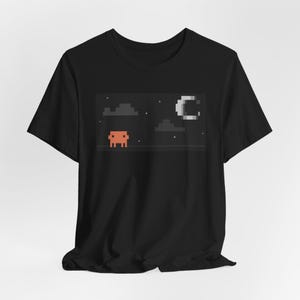 May include: A black short-sleeve t-shirt featuring a pixel art graphic on the chest. The design depicts a small orange square character standing on a dotted line beneath a night sky with grey pixelated clouds, small white stars, and a crescent moon.