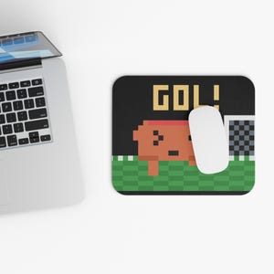 May include: A rectangular mouse pad featuring a pixel art design of a brown character on a green checkered field with a soccer goal and the text GOL! in yellow letters. A white computer mouse rests on the surface of the mouse pad.