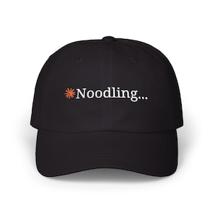 May include: A black baseball cap featuring an embroidered orange starburst icon followed by the white text Noodling... centered on the front panel. The cap has a curved brim and a structured crown with a button at the top center.
