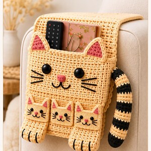May include: A beige crochet sofa armrest organiser featuring a large cat face design with black eyes, a pink nose, and three smaller cat-shaped pockets. It includes a striped black and beige tail, pink-accented ears, and a top compartment.