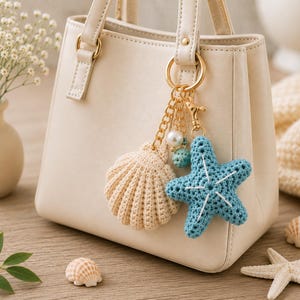 May include: A cream-colored handbag featuring a gold-toned metal keychain with a beige crocheted seashell, a blue crocheted starfish with white stitching, a small white pearl bead, and a tiny textured teal bead attached to the handle.