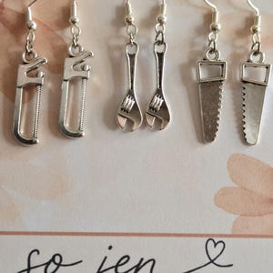 May include: Three pairs of silver-toned metal earrings featuring miniature tool charms: a hacksaw, an adjustable wrench, and a hand saw. Each pair hangs from a silver-toned hook. The display card features the text so jen STUDIOS and #136.