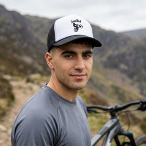 Puede incluir: Una gorra de estilo camionero en blanco y negro con un gráfico de una persona haciendo un caballito en bicicleta y el texto Lean Back impreso sobre la ilustración. La gorra tiene un panel frontal blanco, visera negra y paneles laterales de malla.