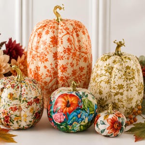 May include: A collection of five decorative pumpkins featuring various floral and botanical patterns in orange, green, and multi-colored designs. Each pumpkin is topped with a metallic gold stem and displays intricate, detailed surface prints.