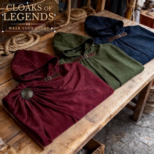 Peut inclure: Trois capes à capuche en bordeaux, vert olive et bleu marine, chacune dotée d'une fermeture à sangle en cuir marron. La cape bordeaux est fixée par une broche métallique circulaire. Le texte CLOAKS OF LEGENDS WEAR YOUR STORY est visible.